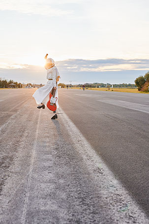 Die Künstlerin Fabiola trägt einen Nonnenkostüm und einen Raumfahrerhelm, während sie mit einer E-Gitarre eine Performance macht. Dabei steht sie in der Abendsonne auf einer leeren Startbahn eines ehem,aligen Flugplatzes